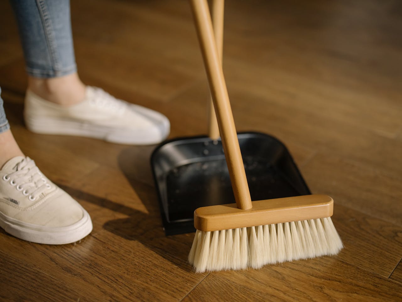 gallery-6 Close-up of cleaning process with broom and dustpan beside sneakers on a wooden indoor floor.
