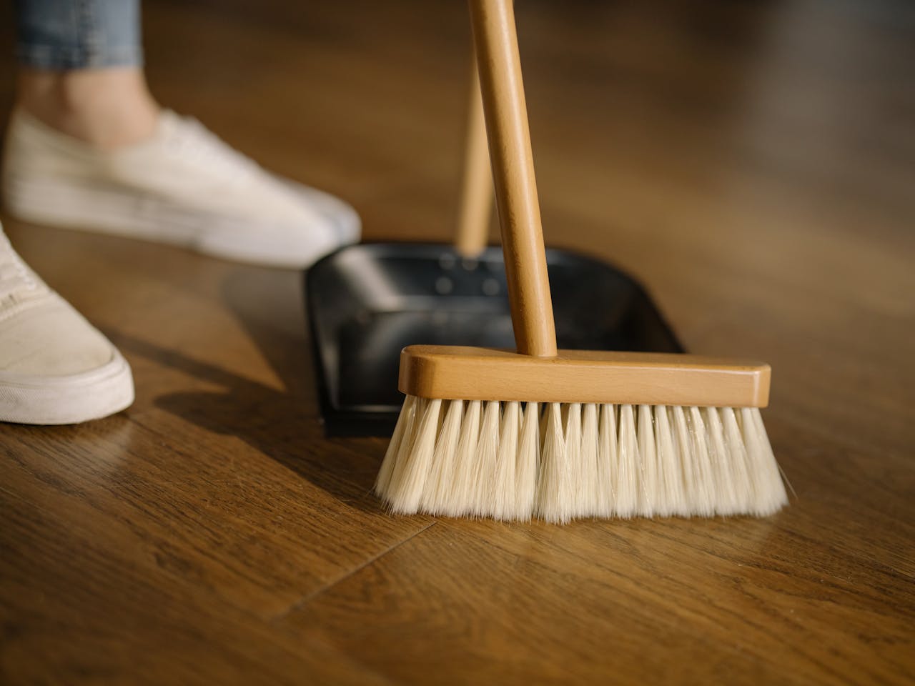 hero-img-02 Close-up of a broom and dustpan with white sneakers indoors, representing housekeeping and cleanliness.